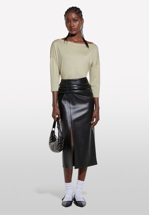Long black leather skirt with a front slit, paired with a loose olive green top. The model carries a black bag with metallic accents.