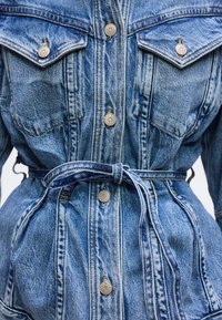 Κοντινό πλάνο μπλε τζιν μπουφάν με μεταλλικά κουμπιά, τσέπες στο στήθος με καπάκια και υφασμάτινη ζώνη δεμένη γύρω από τη μέση.