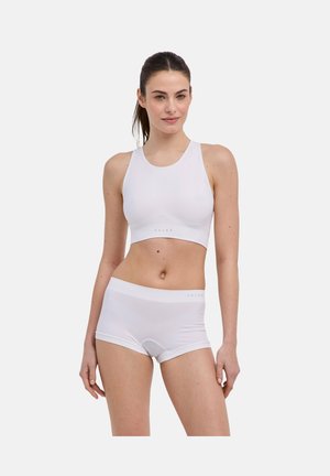 Młoda kobieta stoi w bezszwowym białym sportowym biustonoszu i dopasowanych majtkach typu boyshort na jednolitym tle.