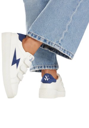 Sneaker bianchi con design a saetta blu scuro e applicazione. Presentano cinturini regolabili e suole spesse e texturizzate. Pantaloni in denim visibili.