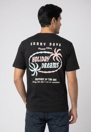 Man facing away wearing a black T-shirt with pastel text and palm tree graphics reading "Sunny Days Holiday Dreams Happiest by the sea."