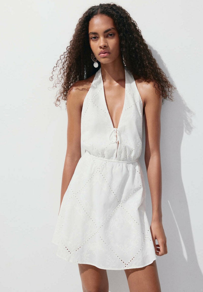 Femme aux longs cheveux bouclés portant une robe blanche sans manches avec un col halter et un motif en losanges, debout devant un fond clair uni.