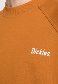 Oranžová mikina vyrobená z texturované tkaniny, s žebrovaným kulatým výstřihem a bílým logem značky "Dickies" potištěným na hrudi.