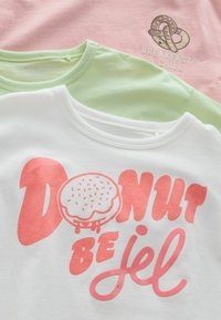 Three t-shirts laid flat: white with pink text and donut graphic; light green; light pink with pretzel and "Breakfast & Chill" text. Soft cotton.