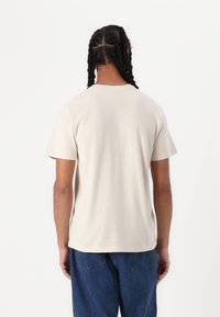 Beige cotton t-shirt with short sleeves, round neckline, and relaxed fit, paired with blue denim pants. Smooth texture, minimal design.