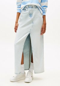 Jupe maxi en denim bleu clair avec une fente avant, dotée d'une taille haute, d'une fermeture par bouton et d'un léger effet dégradé ; associée à des baskets blanches et des chaussettes.