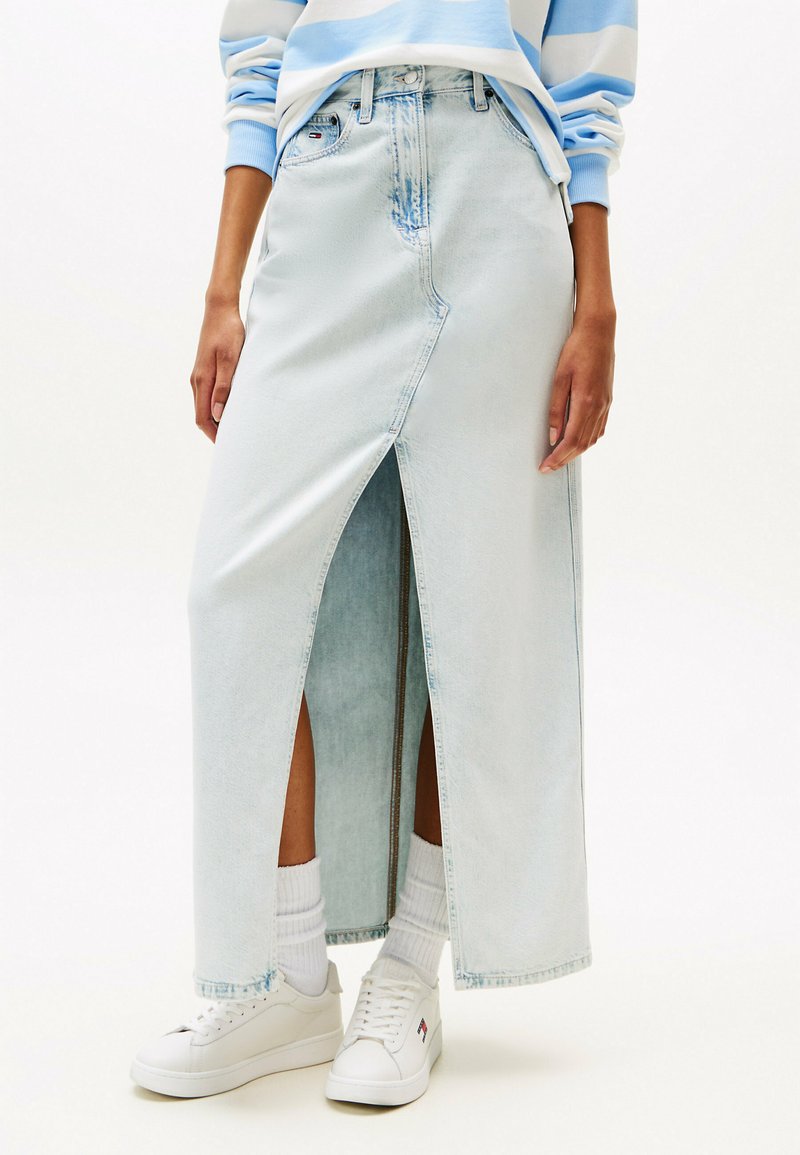 Jupe maxi en denim bleu clair avec une fente avant, dotée d'une taille haute, d'une fermeture par bouton et d'un léger effet dégradé ; associée à des baskets blanches et des chaussettes.