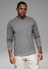 Muž oblečený v šedém tričku s dlouhým rukávem s logem a světlých béžových cargo kalhotách, stojící před jednobarevným šedým pozadím.