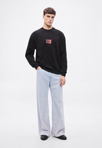 Μαύρο φούτερ με κόκκινο ορθογώνιο λογότυπο που γράφει «HUGO», συνδυασμένο με ανοιχτό μπλε παντελόνι φαρδύ και μαύρα παπούτσια.