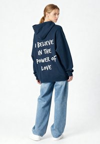 Hoodie marine avec un grand texte blanc dans le dos disant « JE CROIS EN LA PUISSANCE DE L'AMOUR », associé à un jean bleu clair déchiré.