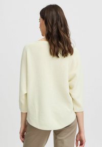 Hellgelbes gestricktes Sweatshirt mit abgerundetem Saum und Dreiviertelärmeln, kombiniert mit hellbeigen Hosen. Weiche Textur und lässige Passform.