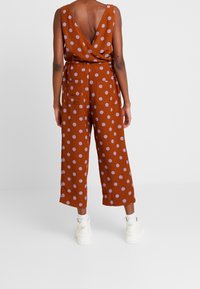 Rdzawoczerwony kombinezon w duże fioletowe kropki, z dekoltem w kształcie V, szerokimi, ściąganymi nogawkami i dwiema tylkami kieszeniami. Wykonany z lekkiej tkaniny.