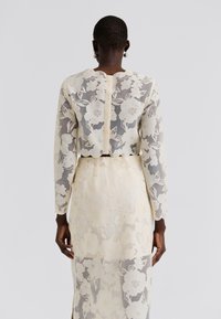 Virágos csipkeruha krém és bézs színekben, hosszú ujjú, testhez simuló felsőrésszel. A áttetsző anyagon bonyolult virágminták díszítik. Hátul cipzáras záródás.