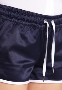 Námořnické modré saténové šortky s elastickým pasem a bílými šňůrkami, s bílým páskem podél lemu, hladkou texturou a uvolněným střihem.