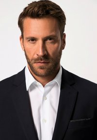 Zwarte blazer met pieklapels over een witte dress shirt. De jas heeft een subtiele accent van een pochet. De stof lijkt glad.
