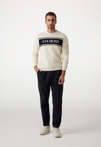 Pulover crem cu dungi orizontale bleumarin, având logo-ul "GUESS". Combinat cu pantaloni închiși la culoare și adidași de culoare deschisă, stând în fața unui fundal simplu.