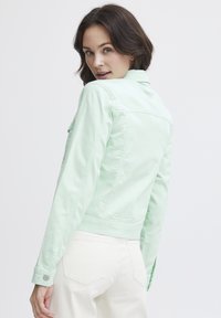 Chaqueta de denim verde menta con un diseño ajustado, puños de botón y detalle en el yugo trasero, combinada con pantalones de color claro.