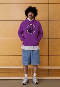 Sweatshirt roxo com capuz e logótipo gráfico, sobreposta a uma camisa branca às riscas, combinada com calções de ganga e sapatilhas desportivas prateadas.
