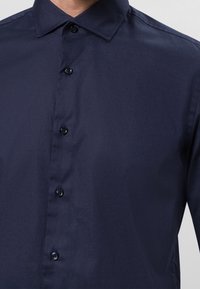 Marinblå knapp-up skjorta, tillverkad av slätt tyg, med en liten krage och sex knappar framtill. Minimalistisk design, inga mönster.