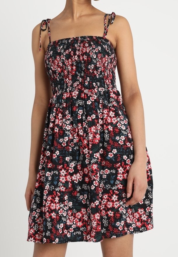 Vrouw in een mouwloos zwart jurkje met een bloemenprint in rood, wit en roze, met geknoopte schouderbandjes.