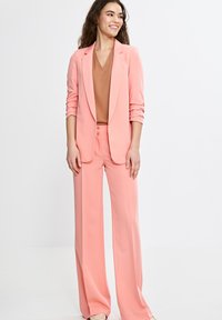 Jasnoróżowy garnitur skrojony na miarę z dopasowaną marynarką i spodniami o szerokich nogawkach. Marynarka ma klapy w kształcie notcha oraz podwinięte rękawy. Pod spodem brązowa bluzka z dekoltem w serek.