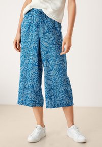 Siniset culotte-housut, joissa on teksturoitu lehtikuvio ja joustava vyötärönauha. Yhdistetty valkoisiin lenkkareihin vaalealla taustalla.