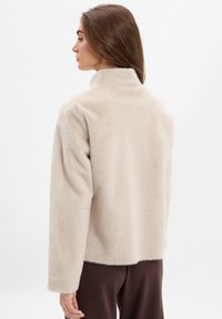 Beige nepbont jasje met een hoge kraag, ronde schouders en een relaxte pasvorm. Zachte textuur en minimaal gestyled.