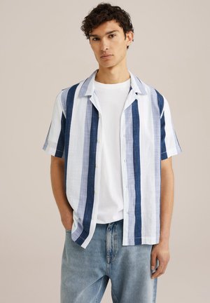 Camisa de manga corta con botones, con rayas verticales en azul claro y oscuro, con un corte relajado, llevada sobre una camiseta blanca. Jeans de mezclilla visibles.