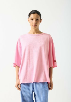 Rozā krāsas pārizmēra T-krekls no mīksta kokvilnas auduma, ar īsām piedurknēm un apaļu kakla izgriezumu. Teksta detaļa kreisajā piedurknē. Kombinēts ar svītrainām biksēm.