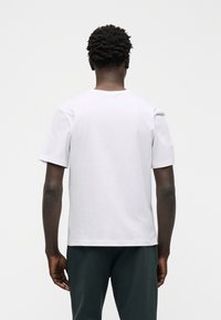 Άνδρας με κοντά, κατσαρά μαλλιά, φορώντας απλό λευκό κοντομάνικο μπλουζάκι και σκουρόχρωμο παντελόνι, στέκεται στραμμένος με την πλάτη σε απλό φόντο.