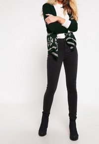 Grön kardiagan med geometriskt mönster, bärs över en vit skjorta. Svarta skinny jeans som kompletterar ankelskor. Mjuka texturer och avslappnad passform.