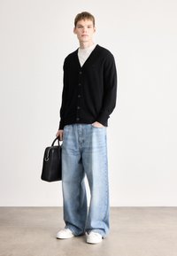 Cardigã preto com botões, combinado com jeans largos azul claro. Ténis brancos e uma mala preta completam o visual. Texturas suaves.