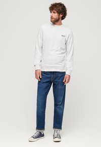 Uomo con capelli ricci e barba, che indossa un felpa grigio chiaro, jeans blu e sneaker alta navy, in piedi davanti a uno sfondo bianco uniforme.