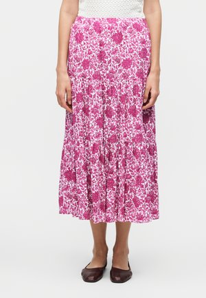 Žena oblečená v ružovej a bielej kvetinovej vrstvenej midi sukni, bielej štruktúrovanej blúzke a tmavohnedých plochých topánkach, stojí pred bielym pozadím.