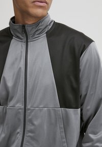 Grå zip-up jakke med høy krage, med svarte detaljer og en blanding av teksturerte paneler, som viser et sporty design.