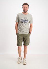 T-shirt grigia con testo stampato blu "Original Apparel," abbinata a pantaloni cargo verde oliva e sneakers beige. Design semplice, outfit casual.
