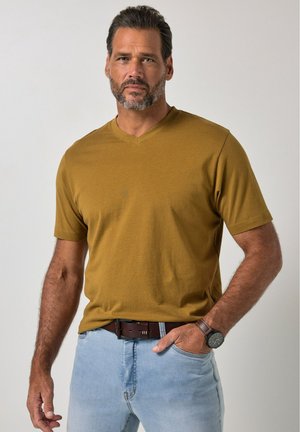 Uomo con barba che indossa una camicia giallo senape a maniche corte, jeans azzurri, cintura marrone e orologio da polso, in piedi davanti a uno sfondo neutro.