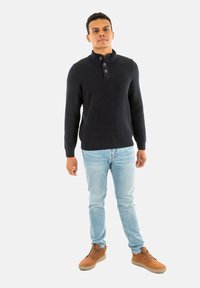 Maglione a coste blu navy con colletto alto e patta con tre bottoni, abbinato a jeans blu chiaro e scarpe stringate marroni.