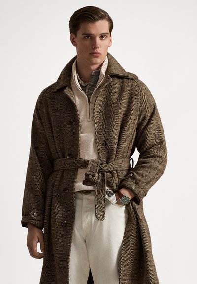 Bruine visgraat woljas met een ceintuur in de taille en grote knopen, gedragen over een beige trui met rits. Lichtgekleurde broek en een horloge zichtbaar.