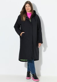 Černý voděodolný kabát s kapucí, plné délky, boční kapsy, spárovaný se zářivě růžovým svetrem a černými džínami, doplněný o růžové masivní boty.