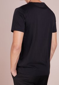 Person, der ein einfaches schwarzes T-Shirt mit kurzen Ärmeln trägt, mit den Händen in den Taschen, von hinten vor einem beigefarbenen Hintergrund gezeigt.