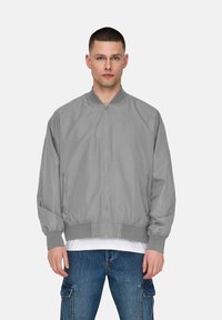 Gray bomber jacket made of lightweight fabric, featuring a zip front, ribbed cuffs, and elastic waistband. Model wears it over a white shirt.
