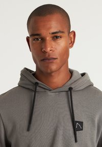 Grijze hoodie met een gestructureerde stof, verstelbare zwarte trekkoorden en een klein zwart logo op de borst.