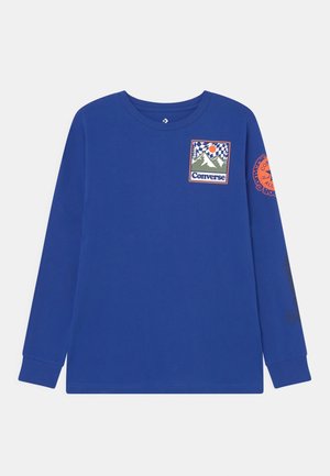 Camisa azul de manga larga con gráfico de montaña y texto "Converse" en el pecho, logo circular naranja en la manga derecha, puños acanalados.