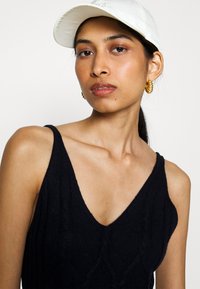 Débardeur noir en tricot avec un décolleté en V profond et une texture côtelée, complété par une casquette blanche de baseball et des boucles d'oreilles en or.