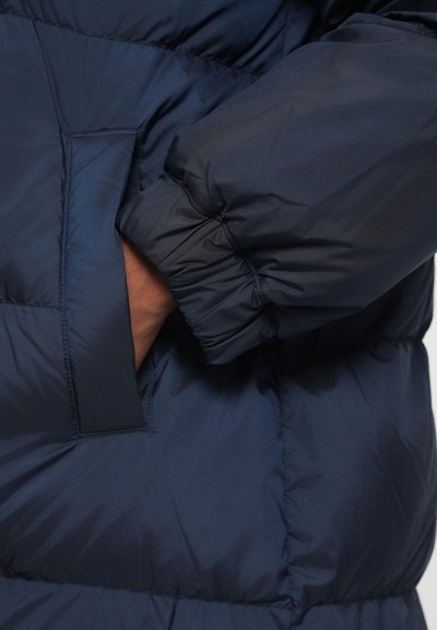 Navy blue puffer jacket featuring a quilted design, visible stitching, elasticated cuffs, and a side pocket with a prominent opening.