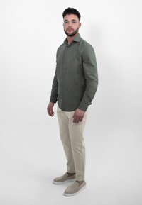 Groene blouse met lange mouwen, met een knoopsluiting aan de voorkant, gecombineerd met beige broeken en lichtgekleurde instapschoenen, staand voor een witte achtergrond.