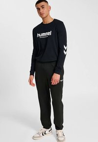 Zwart lang mouw shirt met wit logo en chevron accenten, gecombineerd met zwarte joggers met een logo. Draagt lichte sneakers.