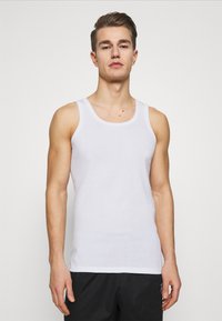 Hvid bomulds tanktop med brede skulderstropper og en rund halsudskæring. Glat tekstur, enkelt design. Båret med sorte shorts.