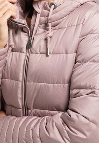 Veste matelassée en mauve clair avec une fermeture éclair, une capuche réglable et des cordons tressés. Présente des coutures horizontales et des accents en métal.